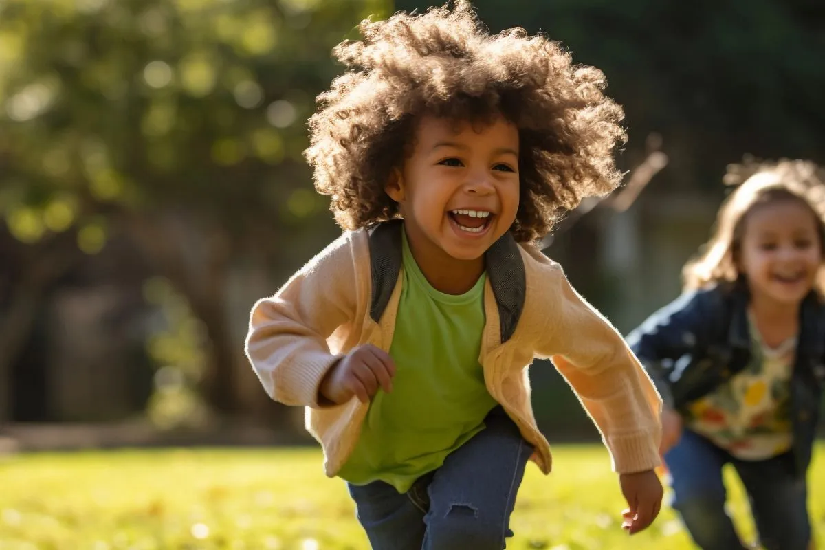 Criança negra com cabelo black power sorri enquanto corre em um parque gramado com outra criança ao fundo.