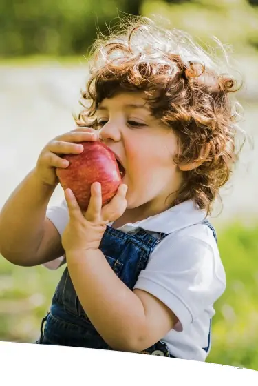 Criança segurando e mordendo uma maçã ao ar livre, representando alimentação saudável para fortalecer a imunidade durante viagens.
