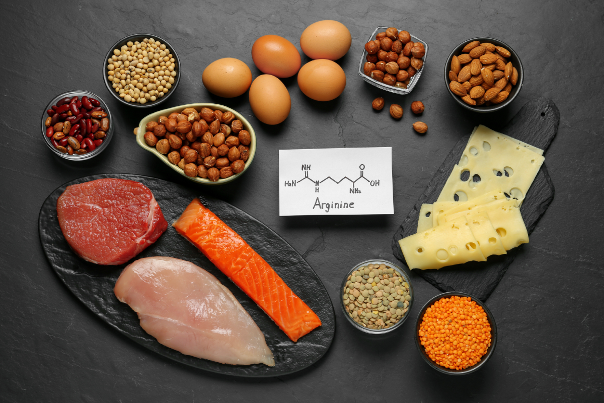 Foto demonstrativa, de uma mesa de pedra preta, com o diagrama químico da arginina, circulado por alimentos que contém o aminoácido: como ovos, queijo, carnes magras, lentilhas e outros.