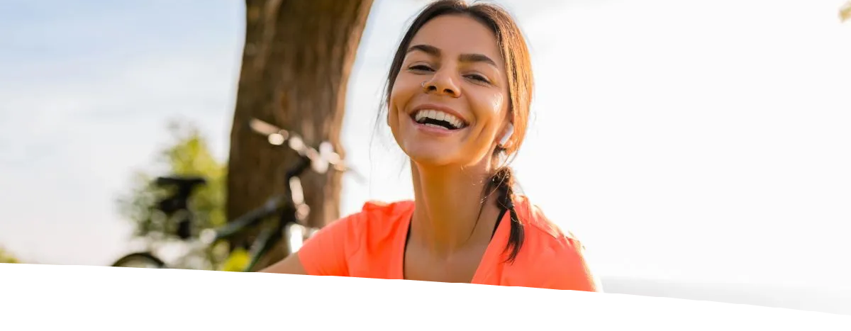 Mulher sorridente sentada na grama de um parque, usando fones de ouvido e segurando um celular, com uma árvore e uma bicicleta ao fundo durante o pôr do sol.