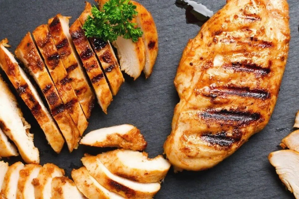 Peito de frango grelhado fatiado em uma tábua escura, decorado com ervas.