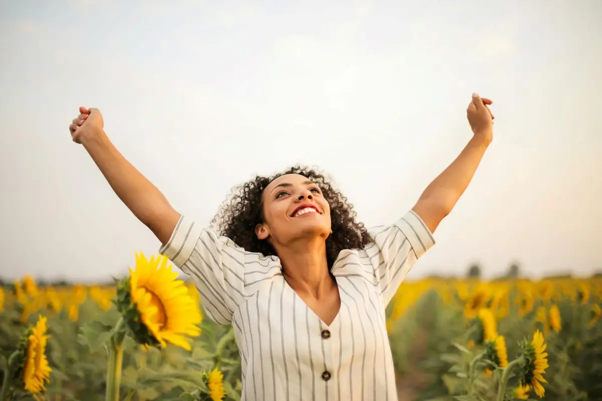Mulher radiante e sorridente com os braços erguidos em um campo de girassóis, transmitindo a sensação de empoderamento, bem-estar e vitalidade. A imagem associa a felicidade e a saúde hormonal feminina com a natureza e a liberdade.