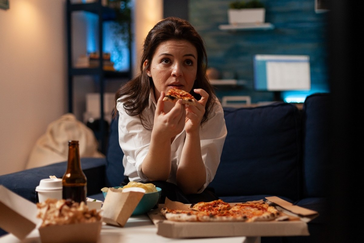 Mulher sentada no sofá, comendo pizza e olhando para a TV, rodeada por fast food e bebidas, em um ambiente de casa.