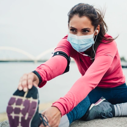 Mulher usando máscara enquanto alonga ao ar livre em um dia frio, representando cuidados e preparação antes do treino.