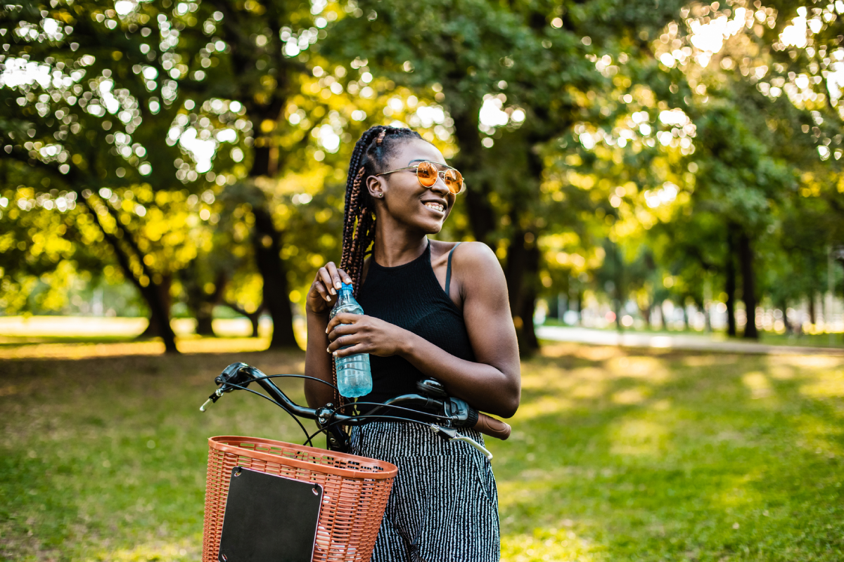Mulher segurando uma garrafinha de água, escorando uma bicicleta, usando óculos escuros, sorrindo em um lindo parque.