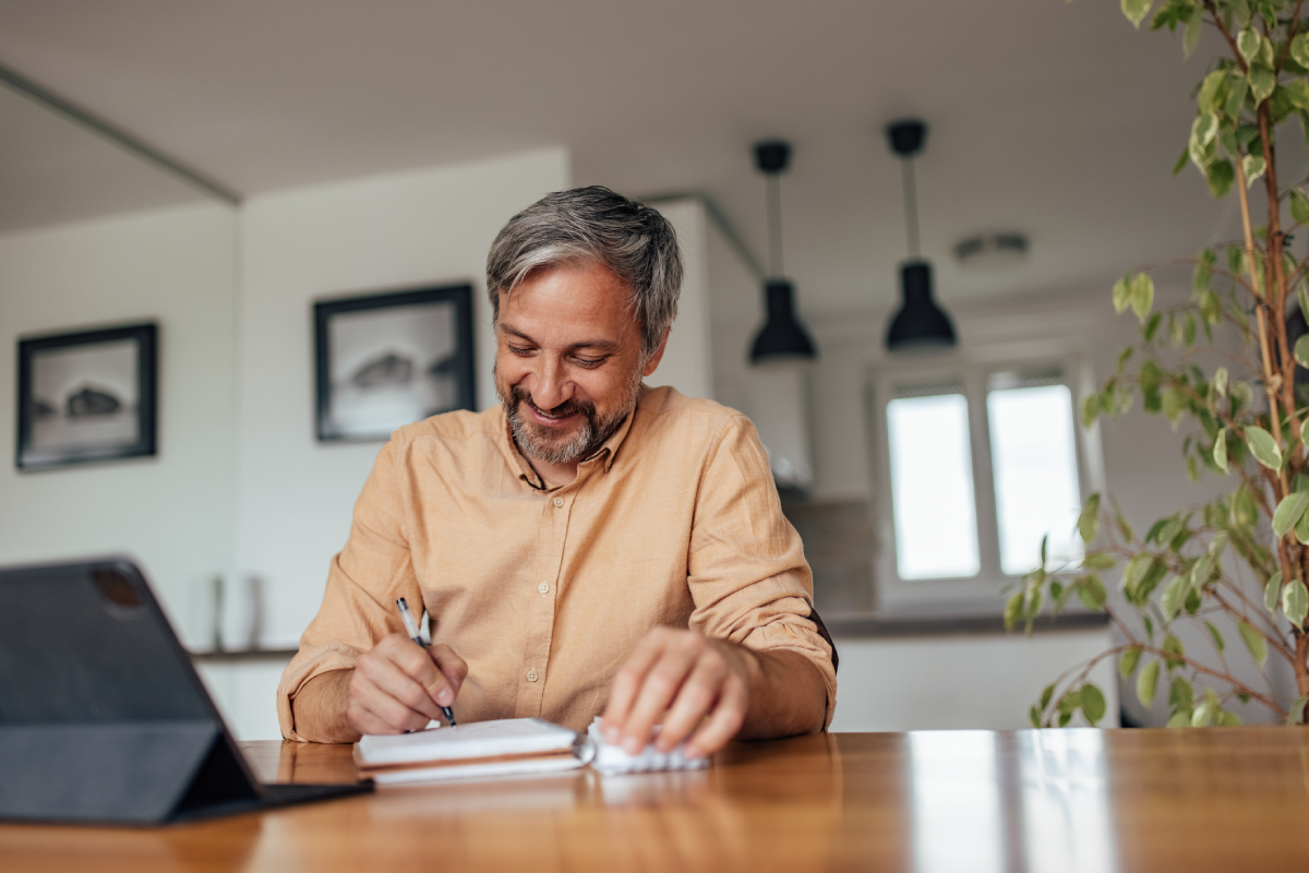 Homem de meia idade fazendo anotações em uma caderna, enquanto assiste algo em seu tablet.