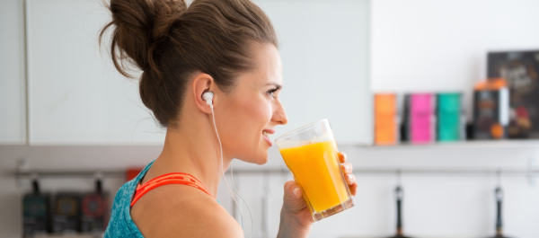 Mulher com coque e roupas leves, usando fones de ouvido, está na cozinha tomando um suco de laranja.