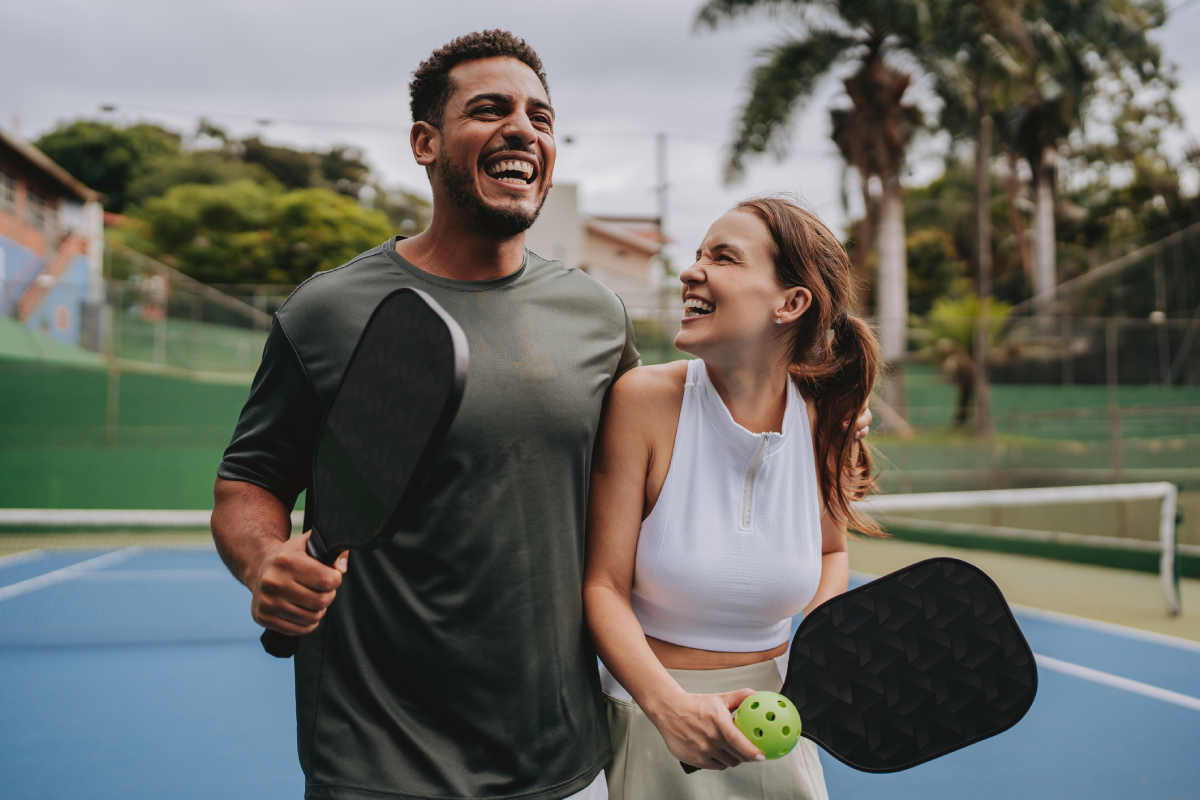 Homem e mulher sorrindo, em quadra, segurando raquetes de pickeball.