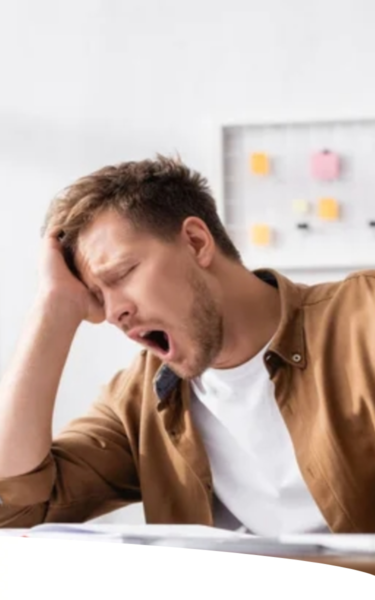 Homem jovem bocejando no trabalho, com expressão de cansaço, sentado em frente ao laptop e segurando a cabeça com uma das mãos.