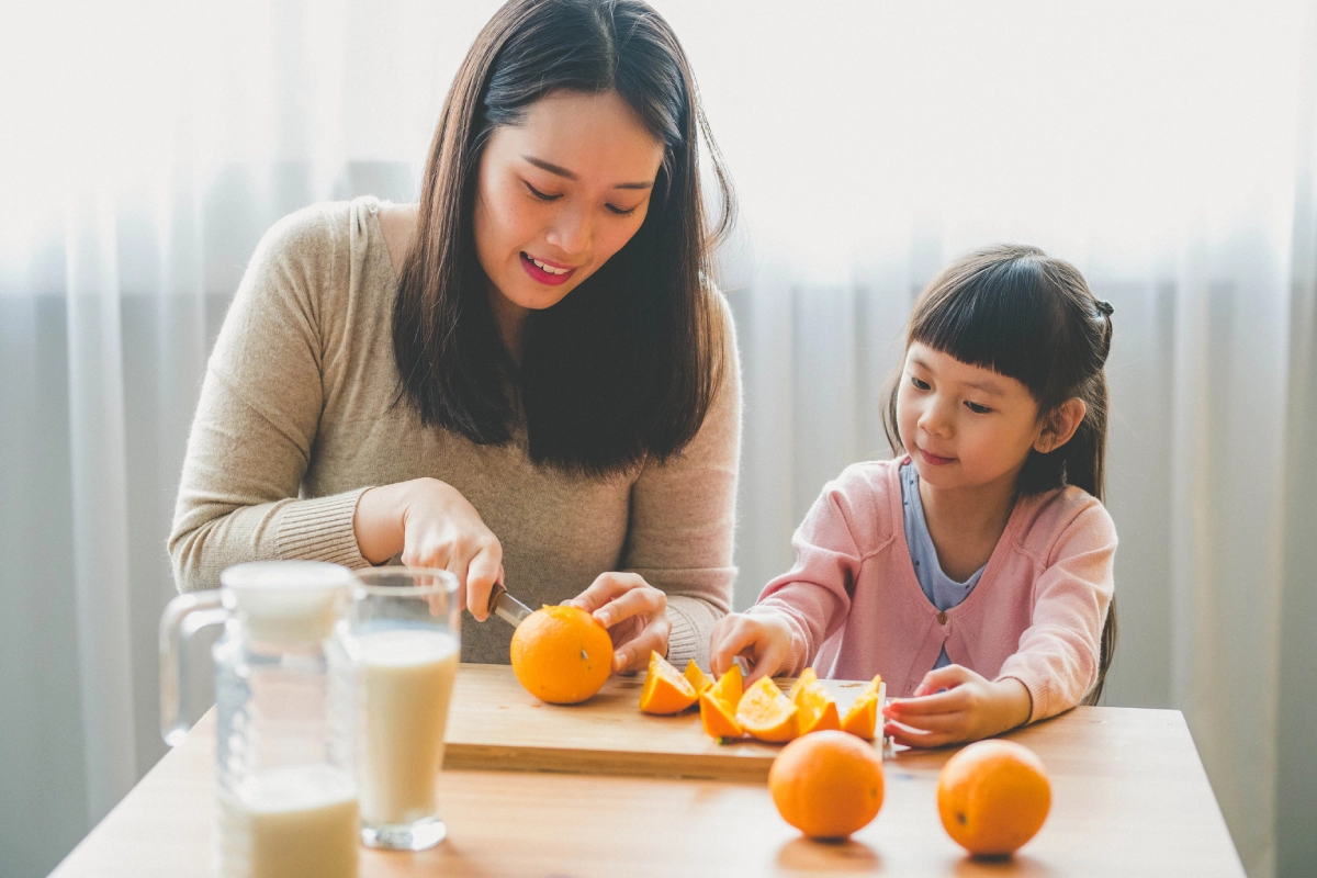 Mãe e filha sentadas à mesa. A mãe está cortando laranjas para a filha.