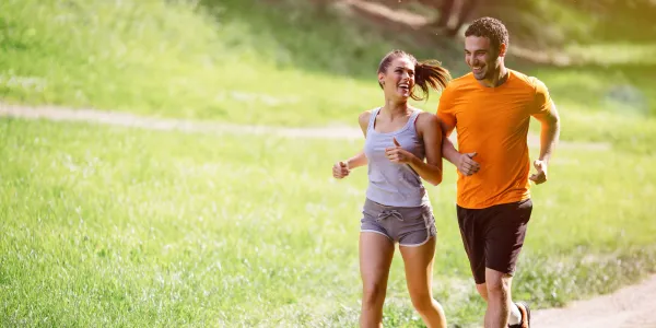 Casal correndo feliz por uma trilha de parque, durante uma tarde ensolarada.
