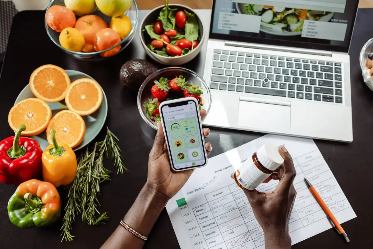Na imagem vemos muitas frutas e verduras, um laptop e um caderno abertos, uma mão segurando vitaminas, outra um celular com app de dieta aberto.