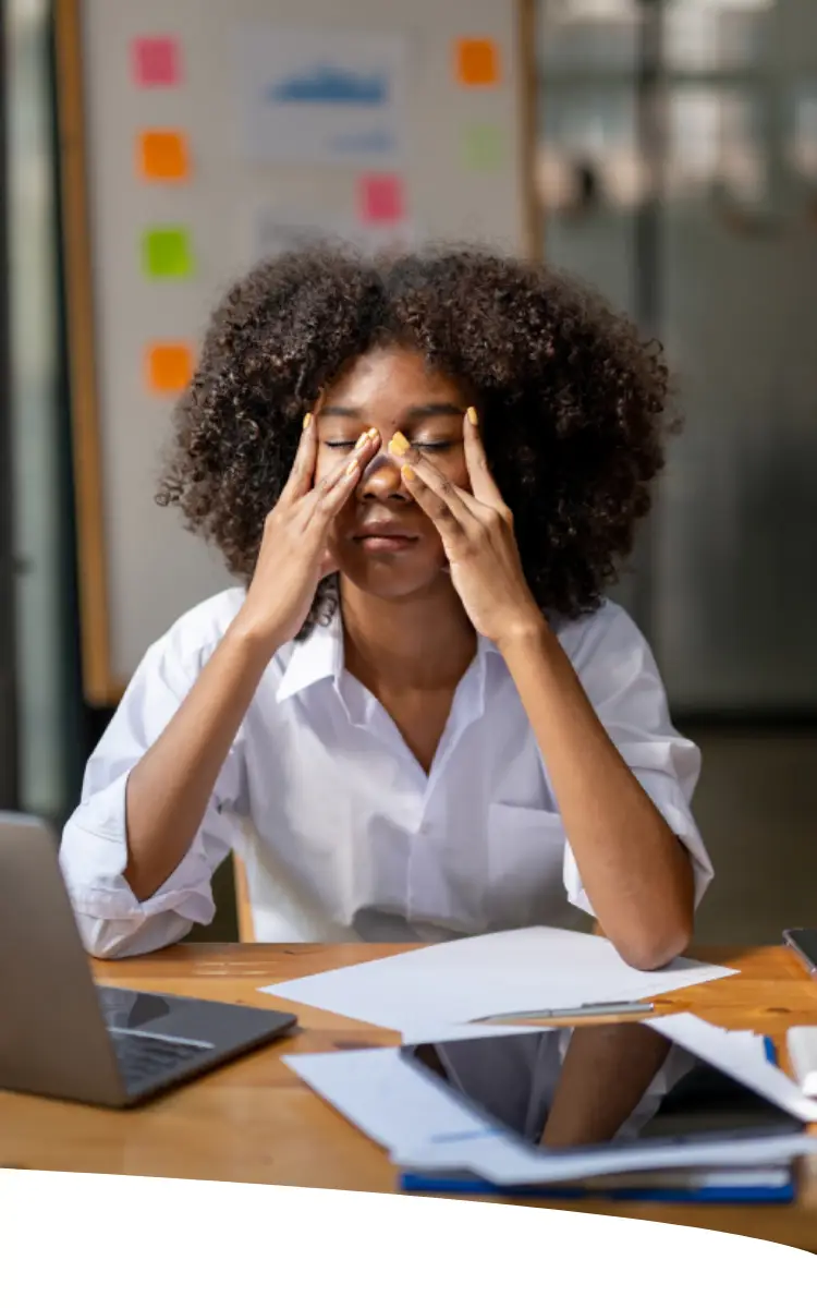 Uma mulher com as mãos no rosto, sentada em sua estação de trabalho, parece estar exausta.