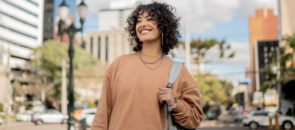 Jovem mulher caminhando em ambiente urbano, sorridente e carregando bolsa.