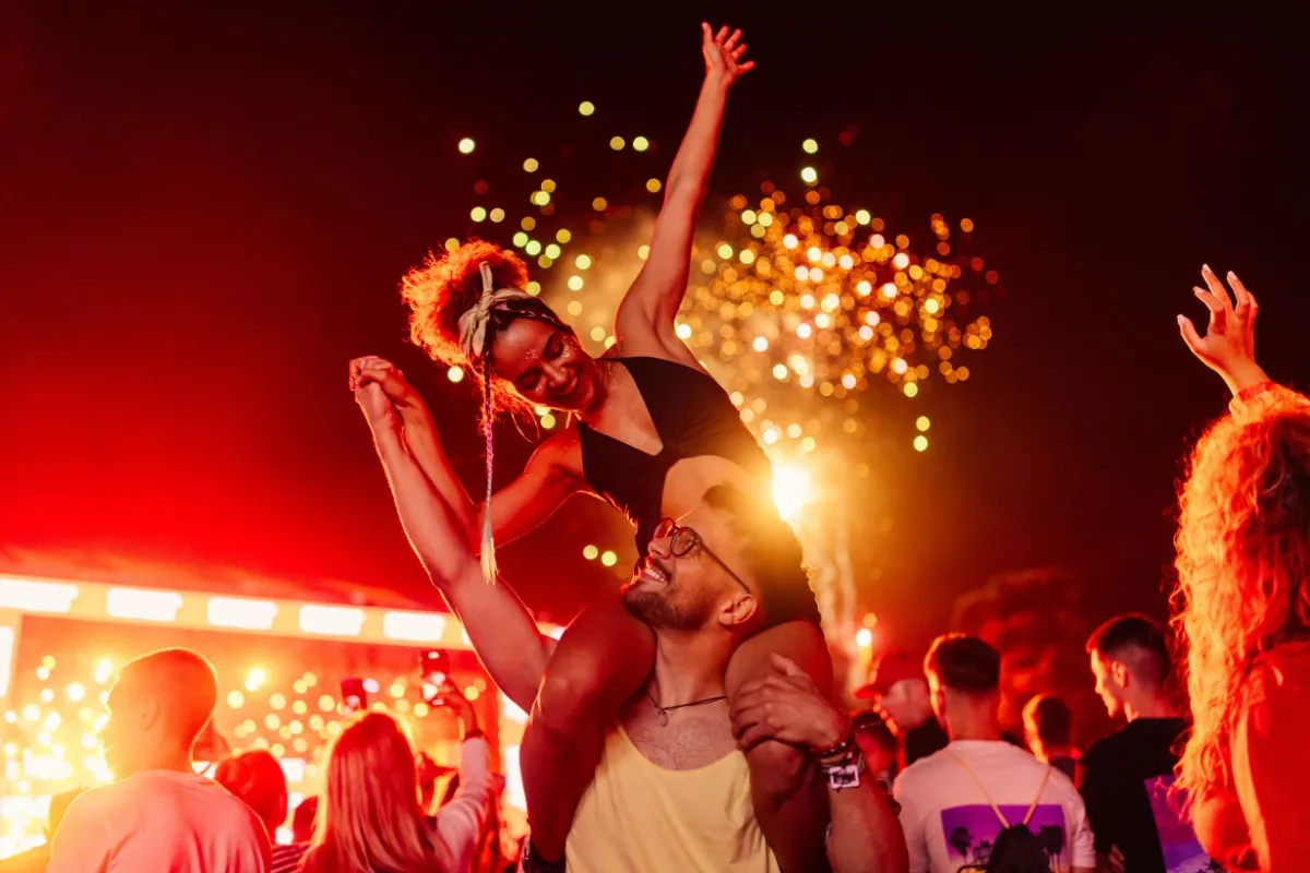 Casal está curtindo um festival de música, à noite, durante queima de fogos. A mulher está sentada nos ombros do homem, segurando a sua mão, enquanto um olha nos olhos do outro.