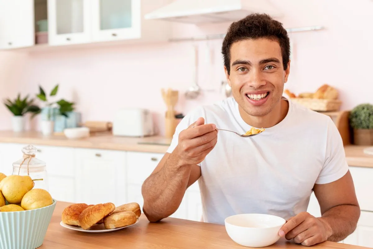 Homem jovem e sorridente, sentado à mesa da cozinha, tomando café da manhã de forma saudável.