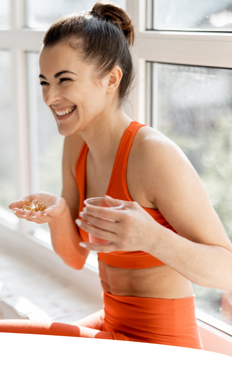Mulher sorrindo, sentada e segurando comprimidos e um copo de água. Ela veste um conjunto laranja e aparece num ambiente majoritariamente branco e com boa iluminação.