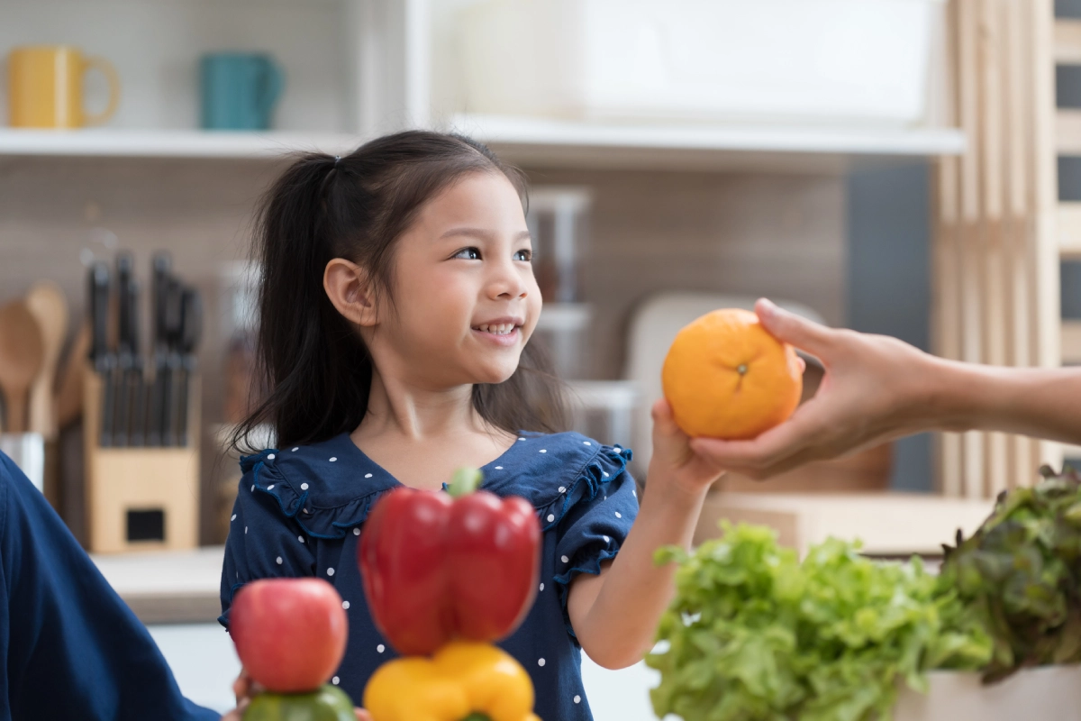 Menina está na cozinha. A mão de um adulto oferece uma laranja para ela, que aceita e sorri, olhando para o adulto (que está fora da imagem).