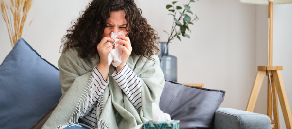 Mulher sentada no sofá, muito agasalhada, assoando o nariz em um lenço de papel.