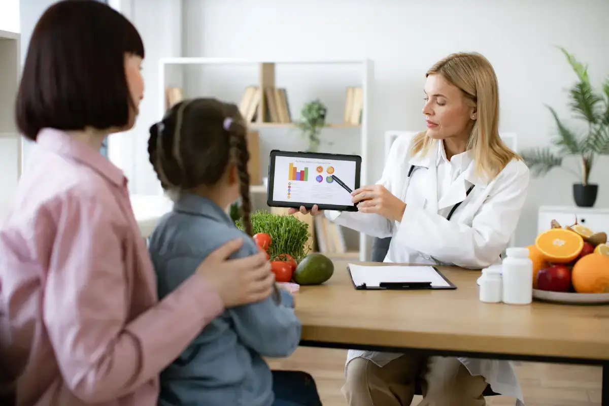 Mãe com criança sentada em seu colo, conversando com nutricionista. Esta aponta dados em seu tablet.