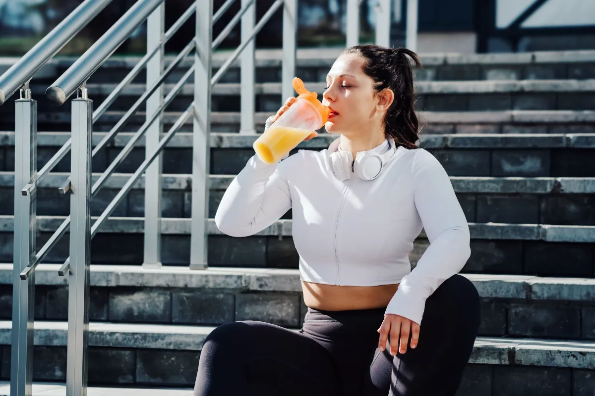 Garota com roupas de academia está sentada em escadaria, enquanto toma um suco de laranja após o treino.