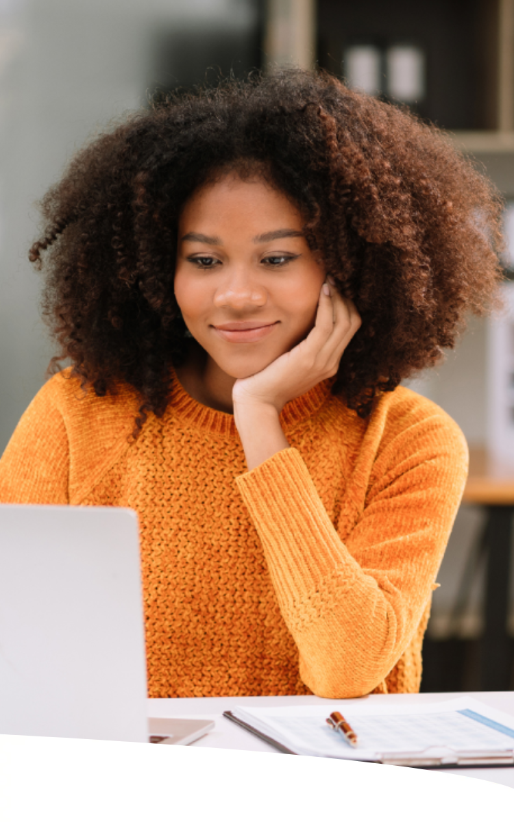 Garota sentada em mesa de escritório, com sorriso no rosto, mão esquerda apoiando o queixo e a outra usando um laptop.