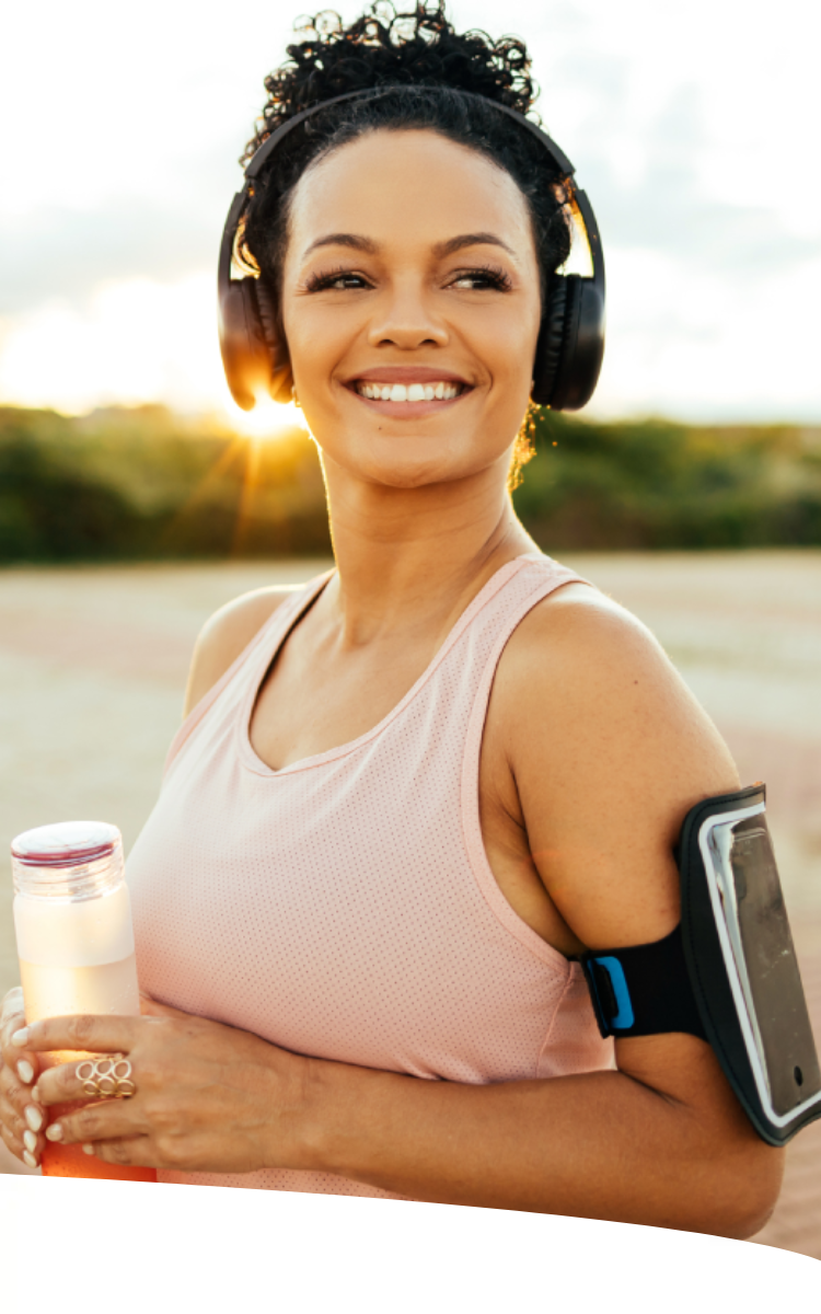 Mulher sorridente, com fone de ouvido, porta-celular no braço e garrafa de chá em mãos, no parque em uma tarde ensolarada.