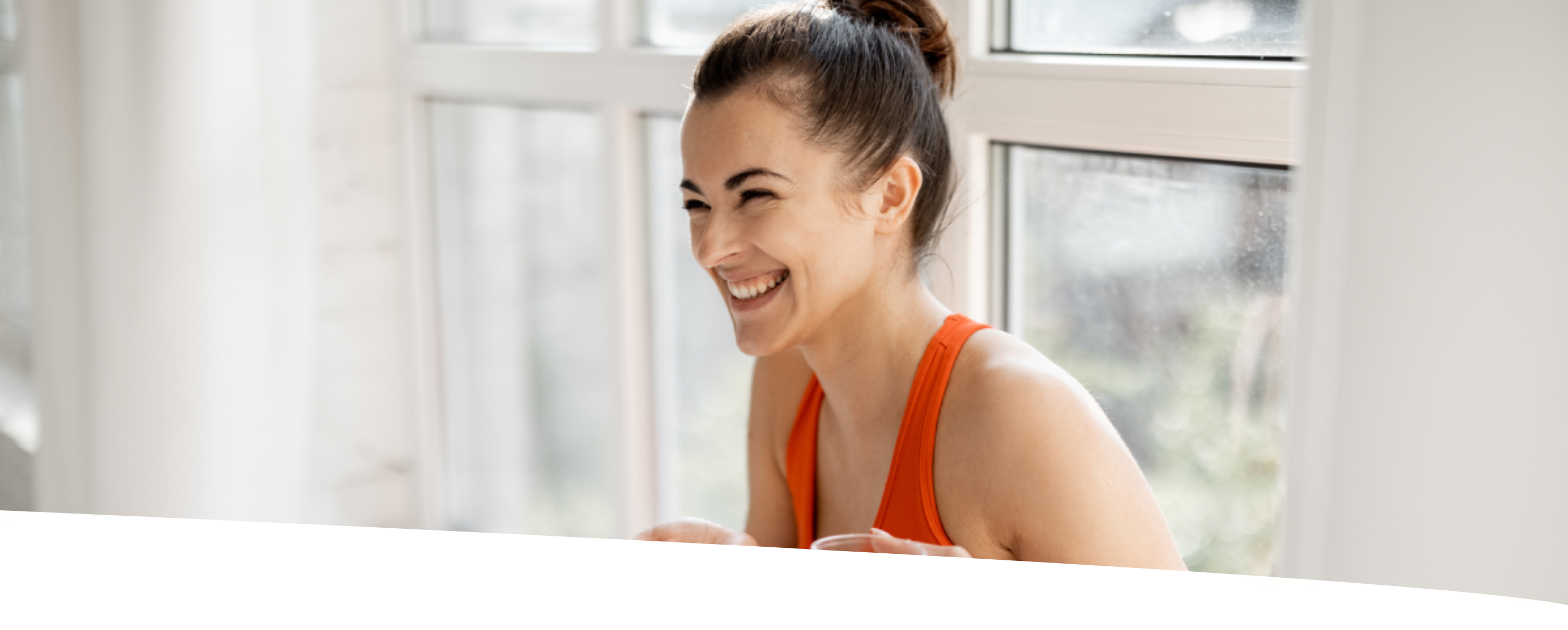 Mulher sorrindo, sentada e segurando comprimidos e um copo de água. Ela veste um conjunto laranja e aparece num ambiente majoritariamente branco e com boa iluminação.