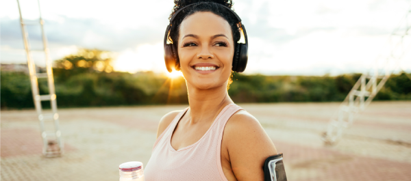 Mulher sorridente, com fone de ouvido, porta-celular no braço e garrafa de chá em mãos, no parque em uma tarde ensolarada.