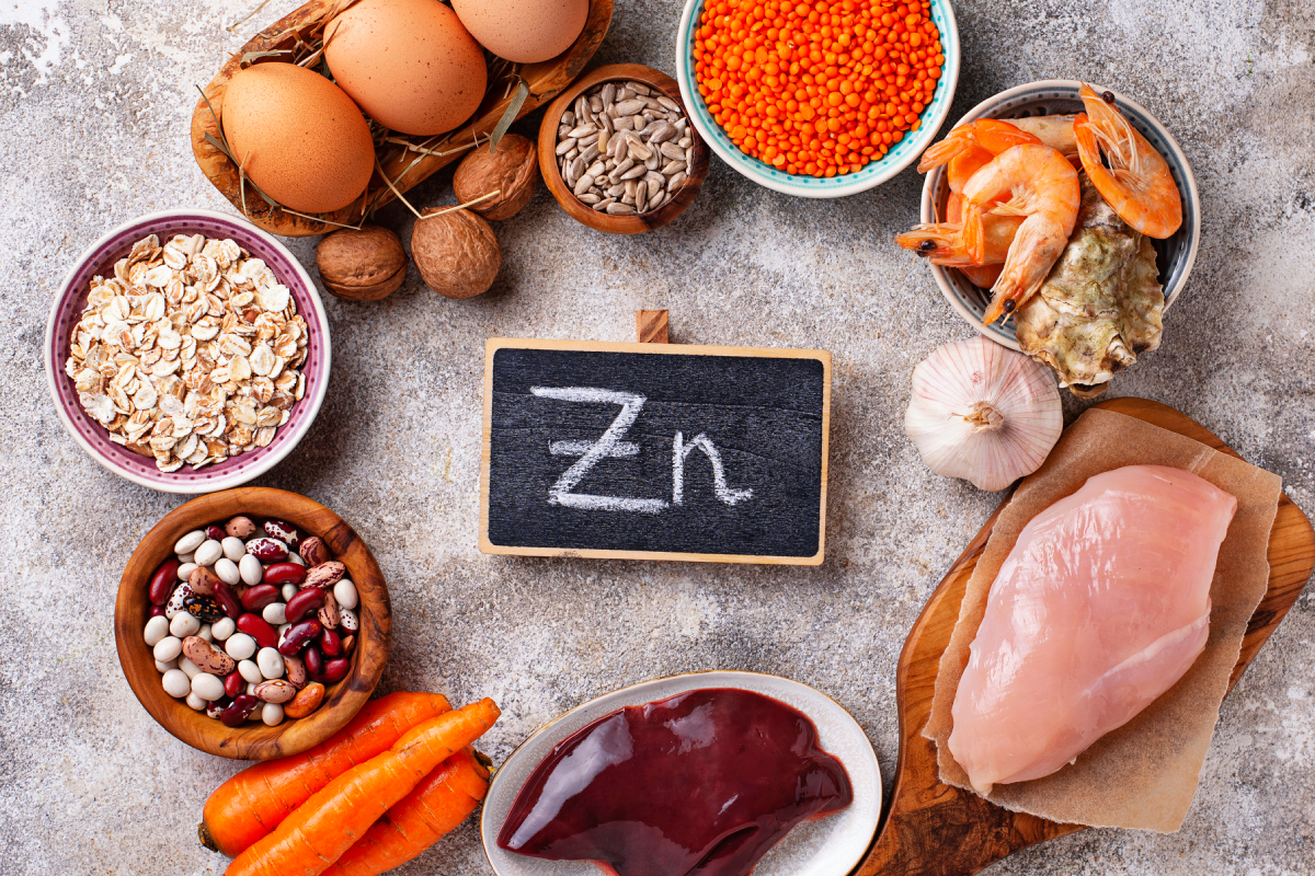 Vários alimentos, como ovos, aveia, leguminosas e cartes, estão dispostos em círculo, com uma placa "Zn" ao meio.