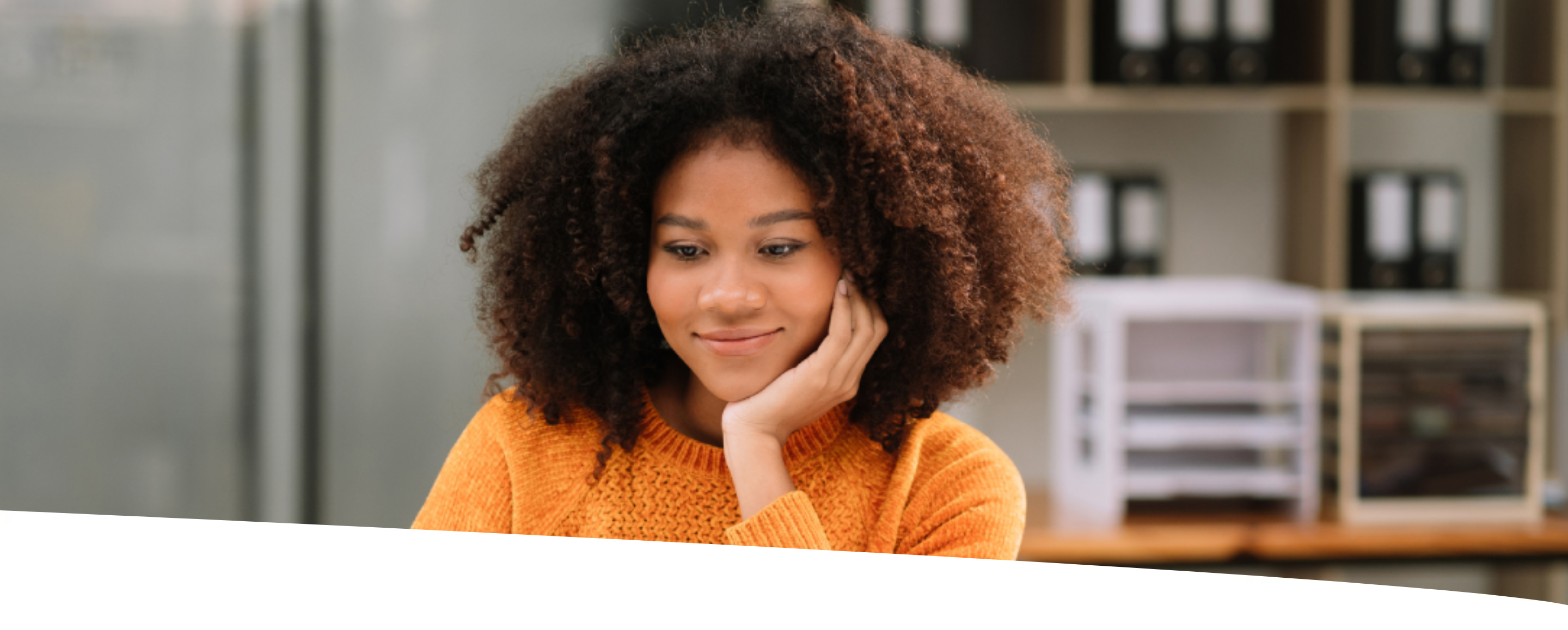 Garota sentada em mesa de escritório, com sorriso no rosto, mão esquerda apoiando o queixo e a outra usando um laptop.