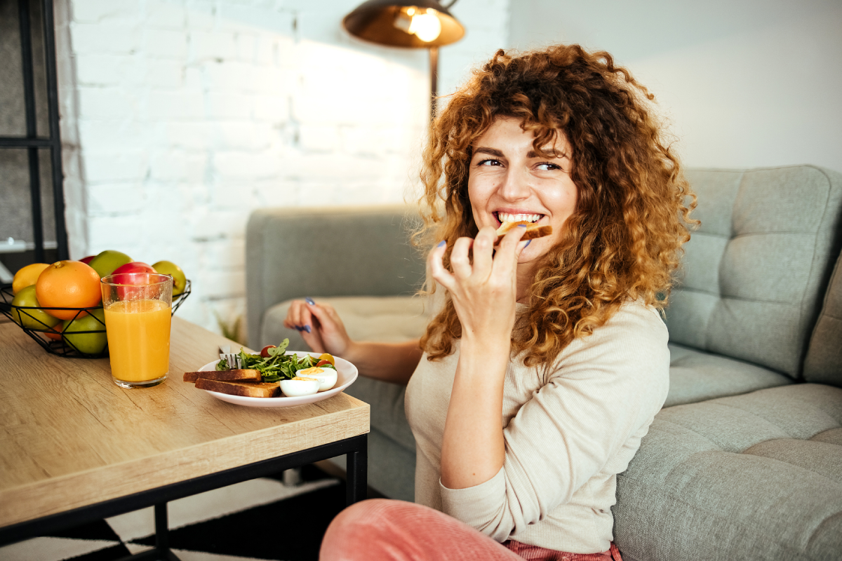 Mulher sorridente comendo salada, sentada no chão de sala de estar.