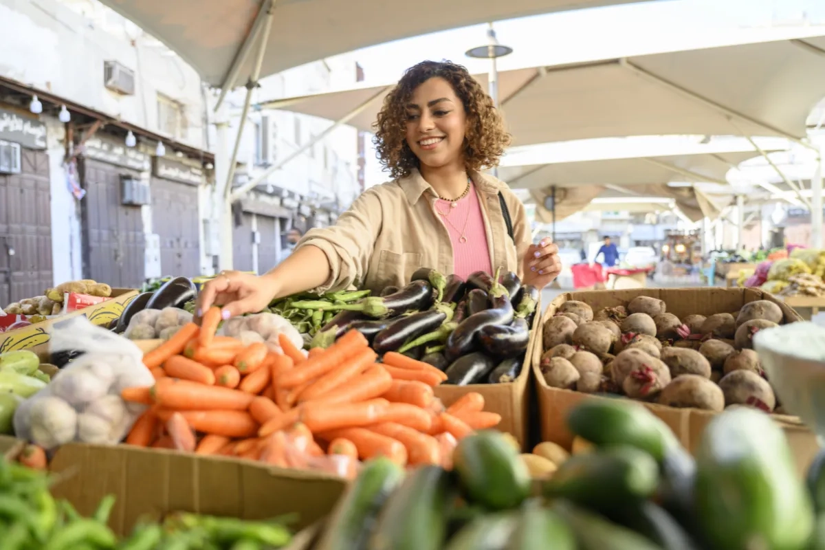 Mulher escolhendo cenouras em uma banca de feira livre. Na banca é possível ver pepinos, berinjelas, alho e outros vegetais.