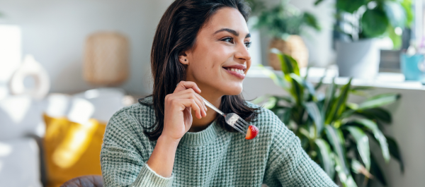 Mulher sorridente comendo um prato de salada com morangos, em uma casa com plantas, olhando para a janela, num dia de sol.