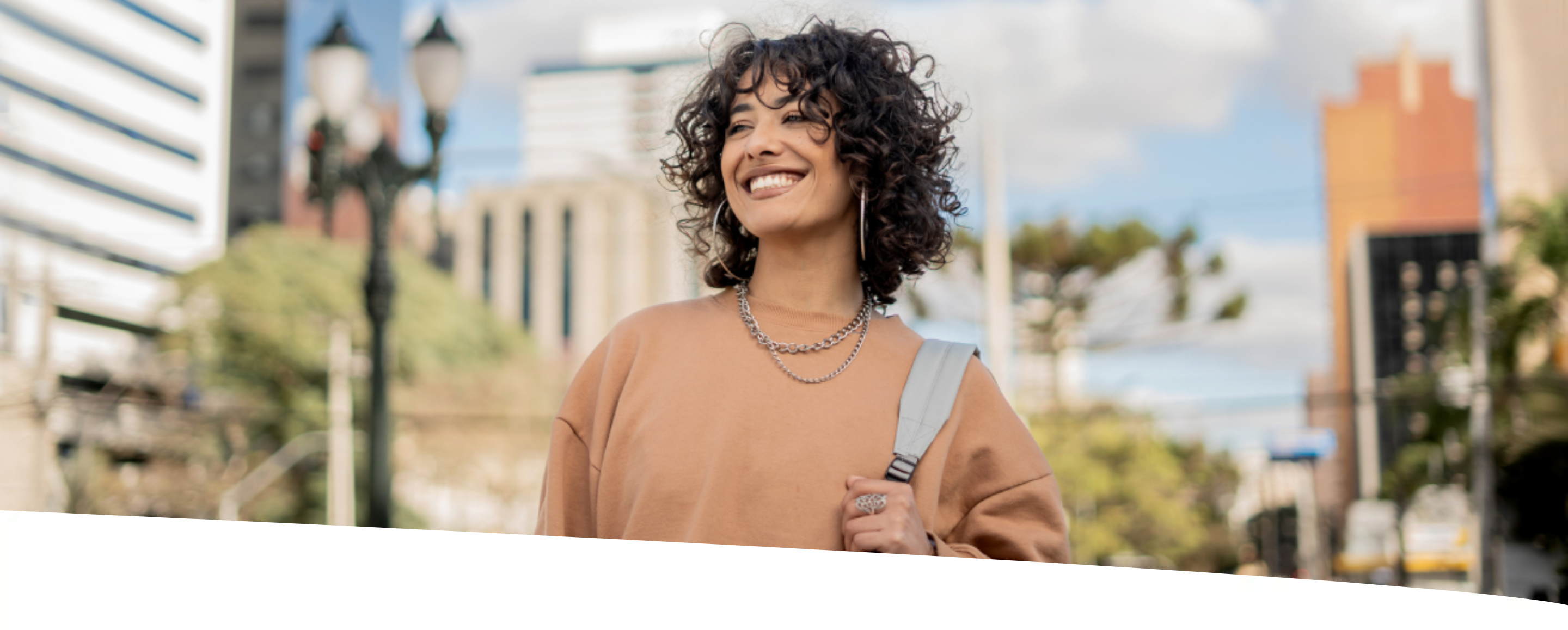Jovem mulher caminhando em ambiente urbano, sorridente e carregando bolsa.