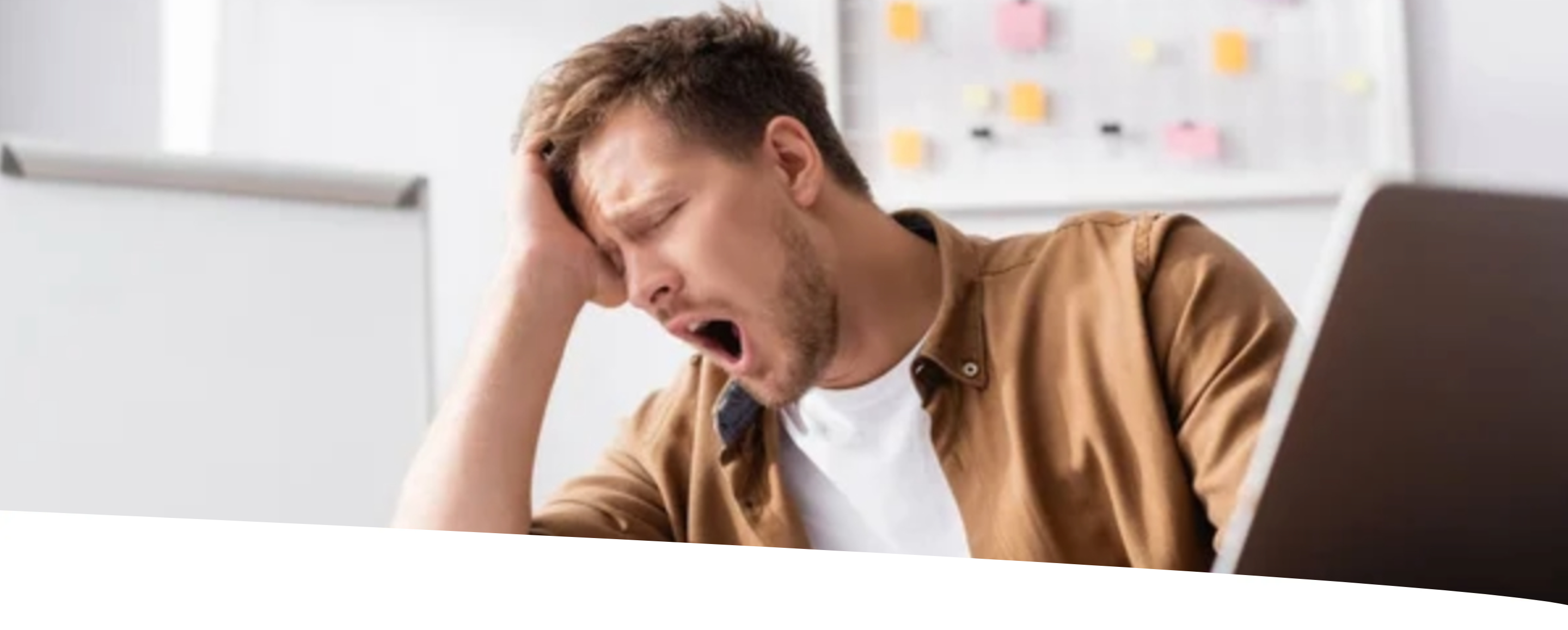 Homem jovem bocejando no trabalho, com expressão de cansaço, sentado em frente ao laptop e segurando a cabeça com uma das mãos.